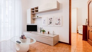 Una sala de estar moderna con una mesa de centro de vidrio, un televisor de pantalla plana, un armario de madera clara y estanterías de pared.