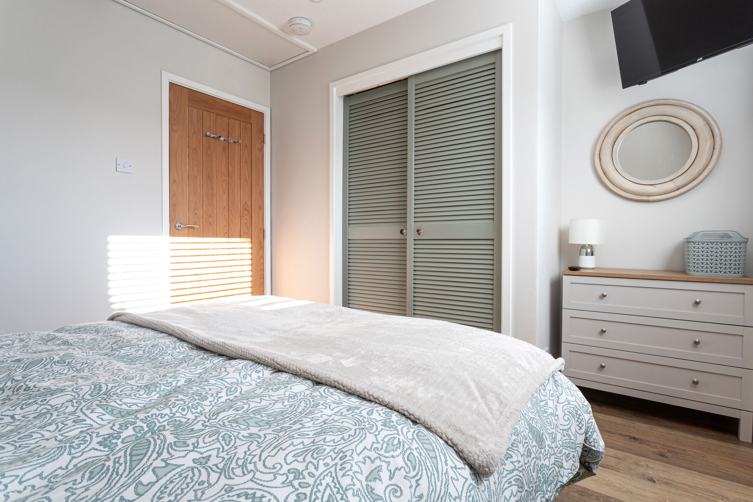 A bedroom with a bed, a wardrobe, a dresser, a mirror, and a television on the wall.