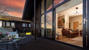 Una terraza de madera con una mesa de comedor y sillas, un sofá, y una cocina visible a través de puertas de cristal.