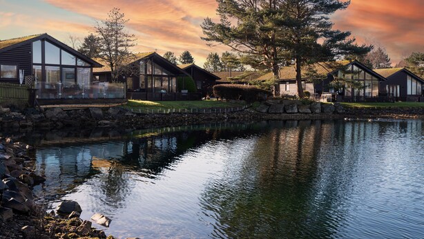 Una cabaña junto al lago con fachada de vidrio, rodeada por un muro de piedra y una terraza de madera.