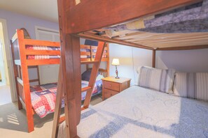 Habitación con literas, una escalera de madera, mesita de noche con lámpara y colcha acolchada.