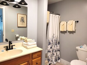 A bathroom with a sink, mirror, and a toilet. There are towels stacked on the counter and a shower curtain.
