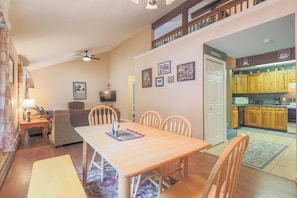 Uma sala de jantar com uma mesa e cadeiras de madeira, uma televisão e uma cozinha ao fundo.