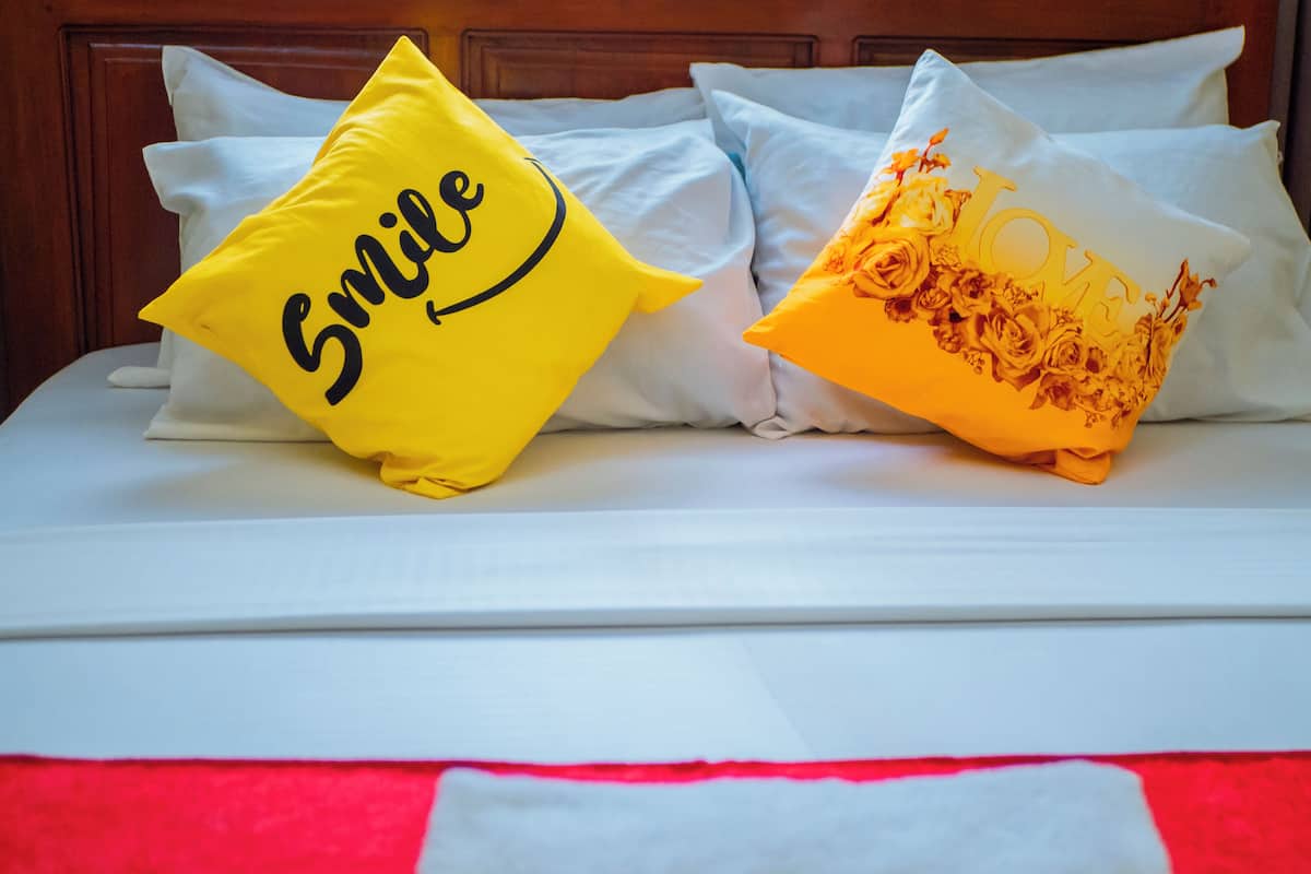 Un lit avec une literie blanche et deux coussins décoratifs, l’un orné d’un visage souriant et l’autre d’un motif floral.