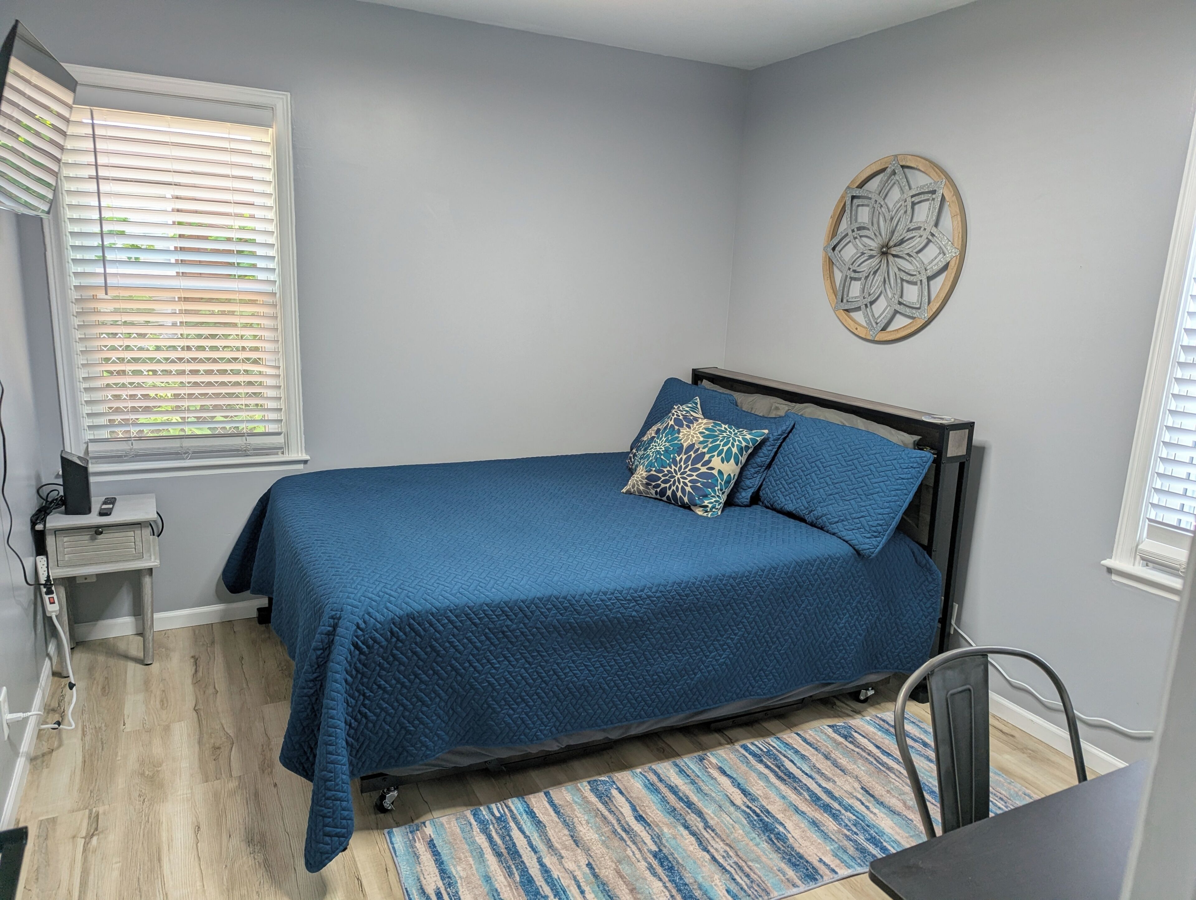 Une chambre à coucher comprenant un lit, une table de chevet, un élément de décoration murale et une fenêtre avec des stores.