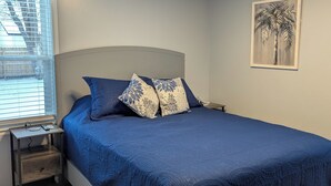 A bedroom with a bed, a nightstand, a lamp, a window with blinds, and a wall art piece.