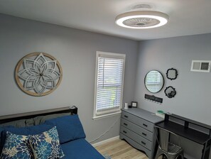 Een slaapkamer met een bed, een bureau, een dressoir, een raam met zonwering, een rond plafondlampje en een decoratie aan de muur.