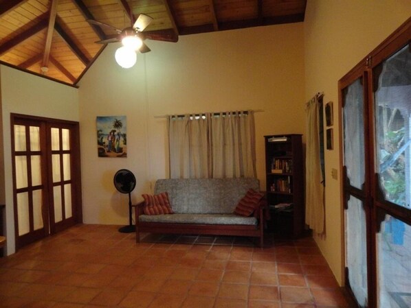 A cozy living room with a wooden ceiling, a sofa, a fan, a bookshelf, and a window with curtains. - Finca Beach House (Placencia)