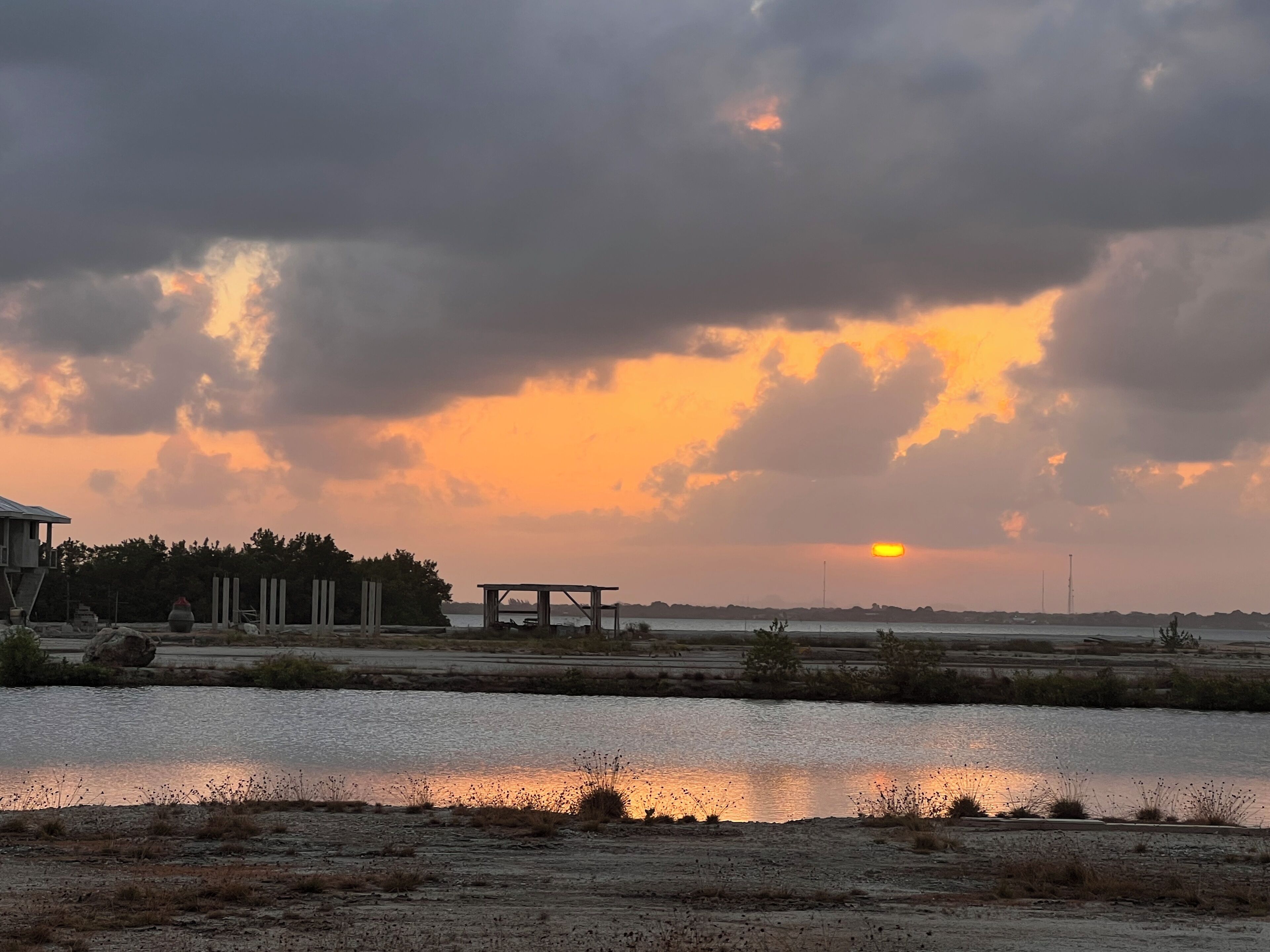 Un coucher de soleil sur une étendue d’eau, avec une structure en bois et des arbres en arrière-plan.