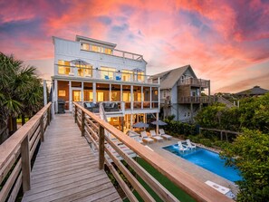 Une propriété en bord de mer, composée de plusieurs étages, dotée d’une terrasse en bois donnant sur un espace piscine, le tout entouré d’une végétation luxuriante.