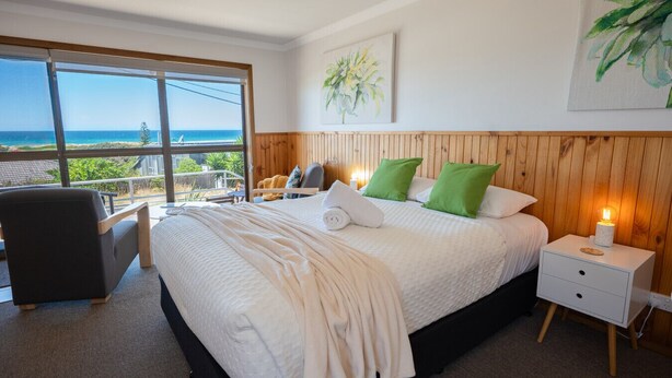 A bedroom with a large bed, a nightstand with a lamp, a chair, and a window with a view of the ocean. - Sea Shells Studio @ The Blue Seas (Scamander)