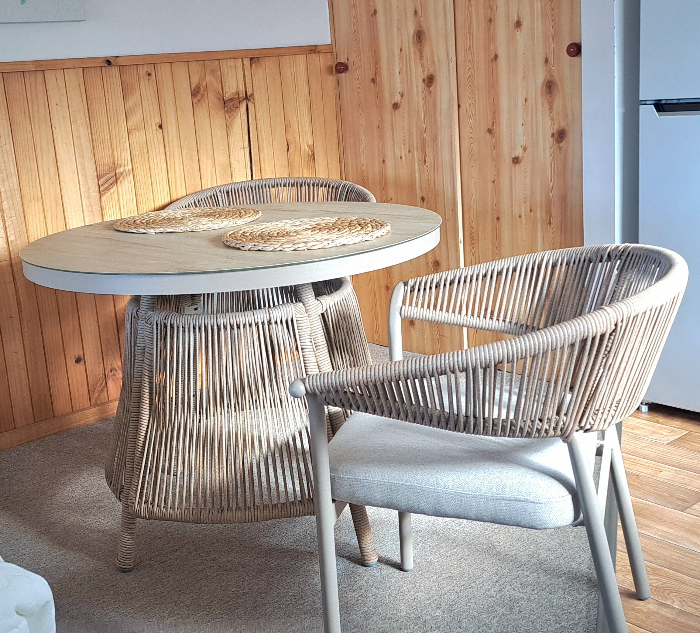 A round glass table with woven placemats, a wicker chair with a cushioned seat, and a wooden wall in the background.