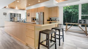 Cocina moderna con armarios de madera, refrigerador de acero inoxidable, microondas y un amplio ventanal con vistas a árboles.