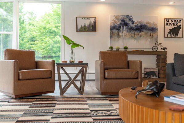 Een woonkamer met twee bruine leren fauteuils, een houten salontafel en een tapijt met een patroon. Op de muur hangen ingelijste foto's en op een bijzettafeltje staat een klein plantje.