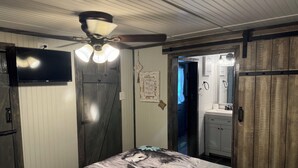 A bedroom with a bed featuring a large, detailed artwork, a ceiling fan, a television mounted on the wall, and a bathroom visible through an open door. - Peaceful cabin retreat to enjoy all the quiet & beauty nature has to offer. (Mansfield)