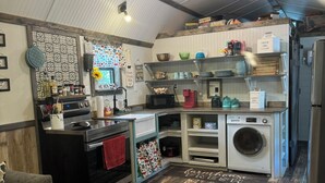 A compact kitchen with a stove, oven, microwave, and a washing machine. There are shelves with various kitchen items and a red towel hanging on the oven. - Peaceful cabin retreat to enjoy all the quiet & beauty nature has to offer. (Mansfield)