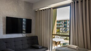 A modern hotel room with a large flat-screen TV mounted on the wall, a dark grey sofa, a small bed with a grey and white striped bedspread, and a large window with curtains. - Mana Beach Experience B 114 Muro Alto (Ipojuca)
