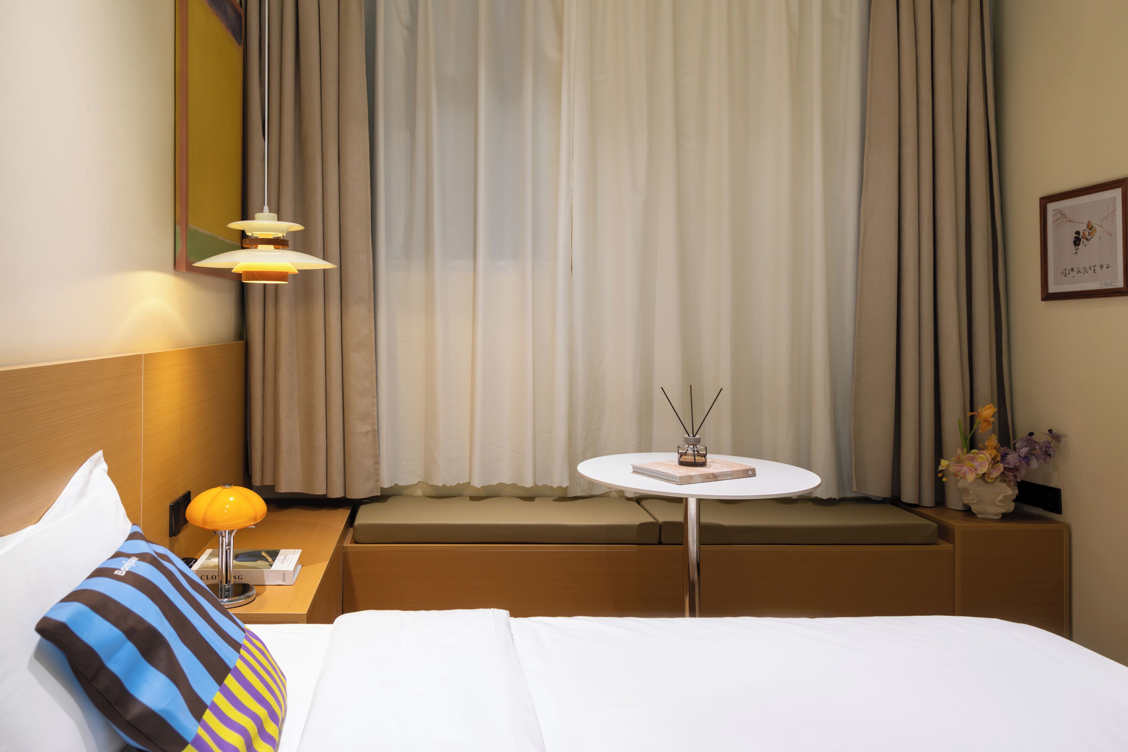 A hotel room with a bed, a bedside table with a lamp, a small round table with a book and a vase of flowers, a window with curtains, and a framed picture on the wall.