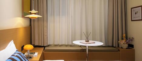 A hotel room with a bed, a bedside table with a lamp, a small round table with a book and a vase of flowers, a window with curtains, and a framed picture on the wall.