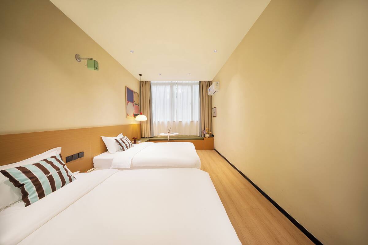 Une chambre d’hôtel avec deux lits, une fenêtre avec des rideaux, une table de chevet et un climatiseur fixé au mur.
