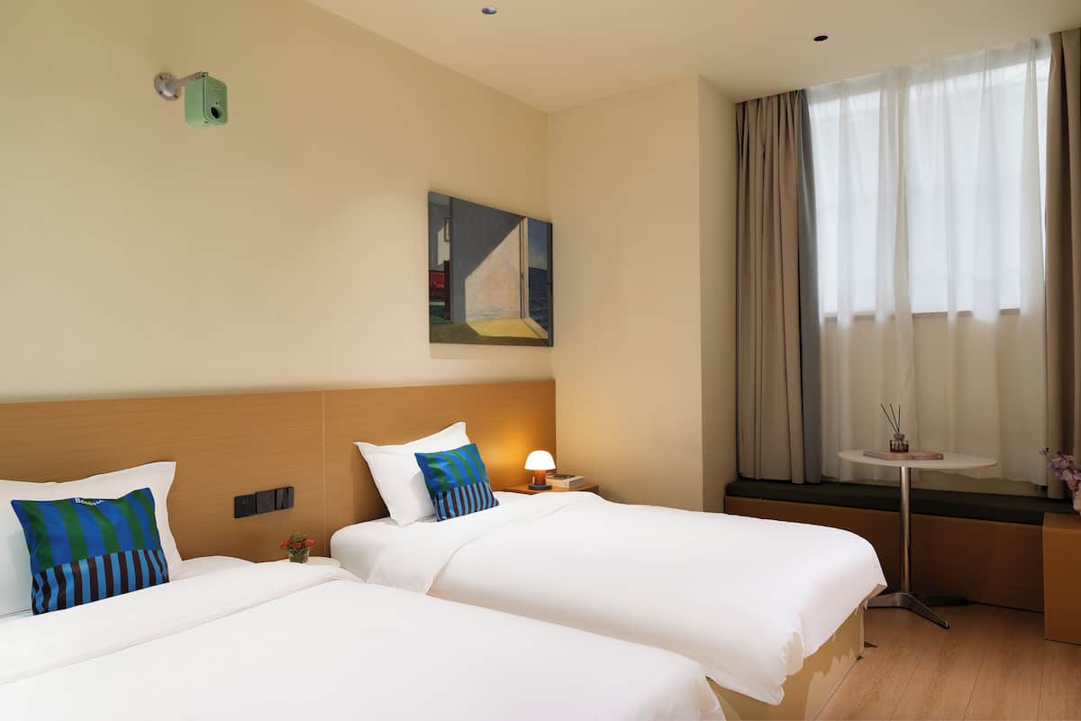 Une chambre d’hôtel avec deux lits, un tableau au mur, une petite table avec une lampe et une fenêtre avec des rideaux.