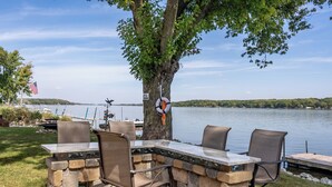 Un espacio de comedor al aire libre con mesas y sillas de piedra, con vistas a un lago con un muelle y una bandera.