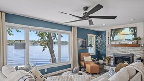 Una sala de estar con un ventanal que da a un lago, una chimenea y un ventilador de techo.