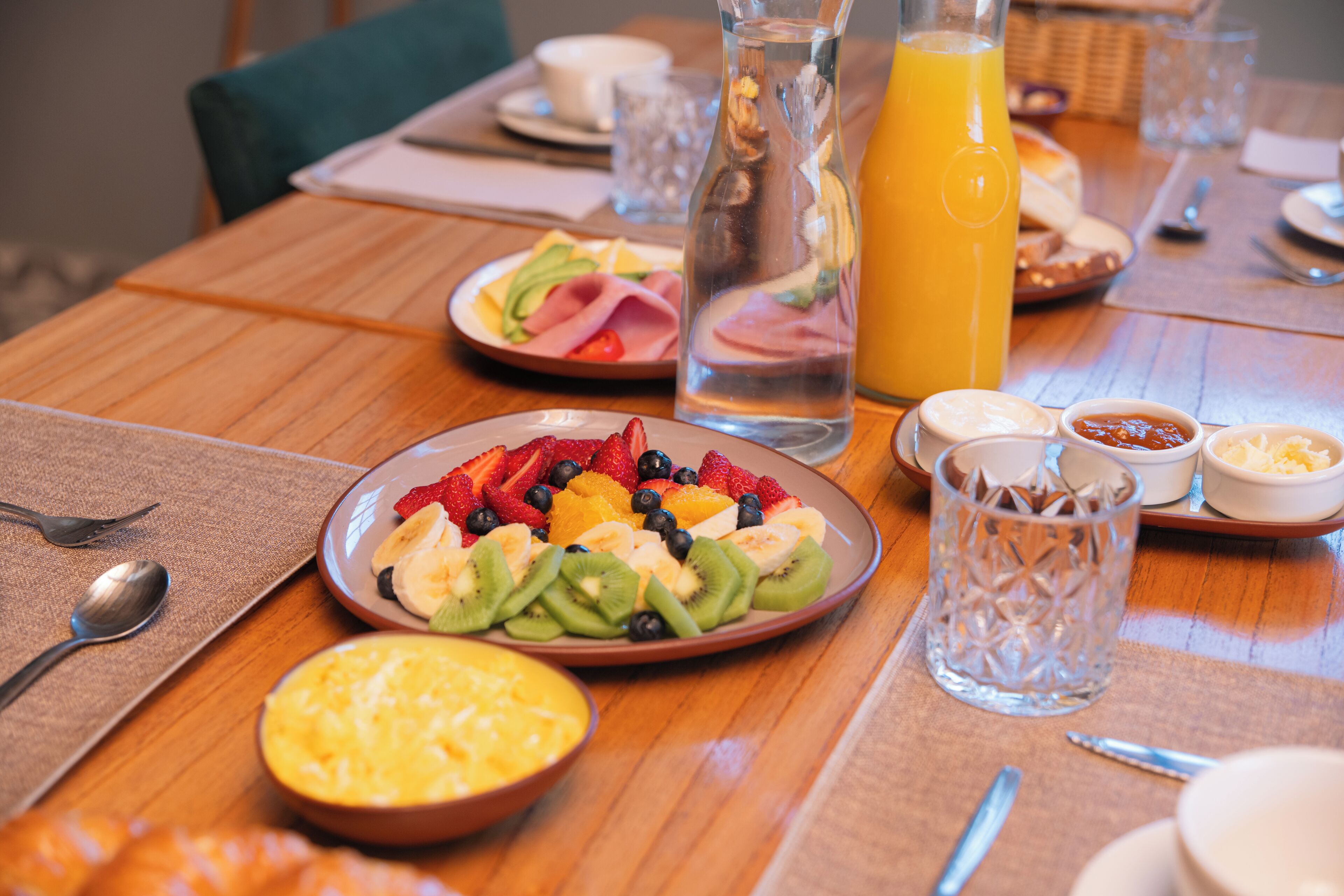 A breakfast setting with plates of fruit, a glass of orange juice, and various condiments on a wooden table.