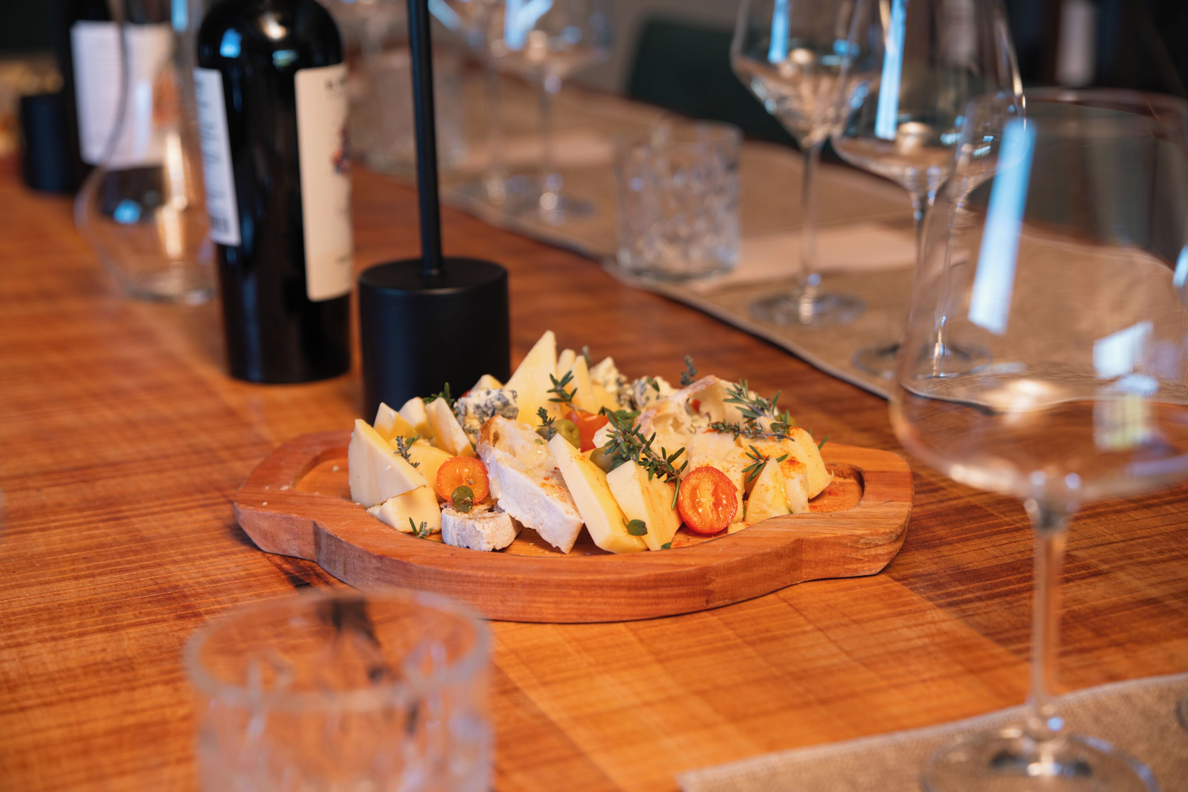 Uma tábua de madeira com uma salada, copos de vinho e uma garrafa sobre uma mesa.