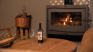 Une pièce chaleureuse avec une cheminée, une table en bois sur laquelle se trouve une bouteille de vin et deux verres, et un panier en osier en arrière-plan.