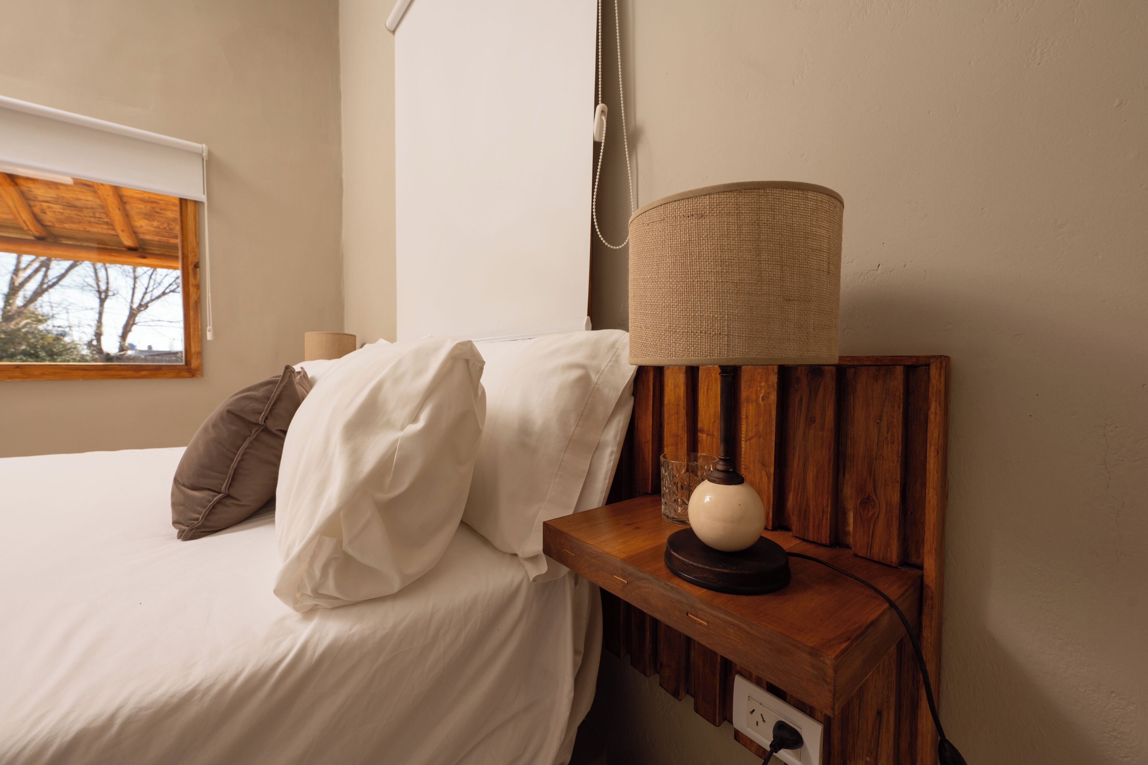 A neatly made bed with white and brown bedding, a wooden bedside table with a lamp, and a window with a view of trees.
