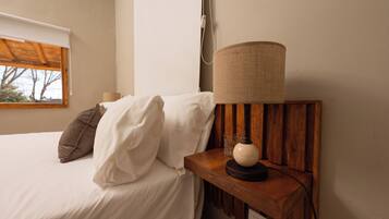 A neatly made bed with white and brown bedding, a wooden bedside table with a lamp, and a window with a view of trees.