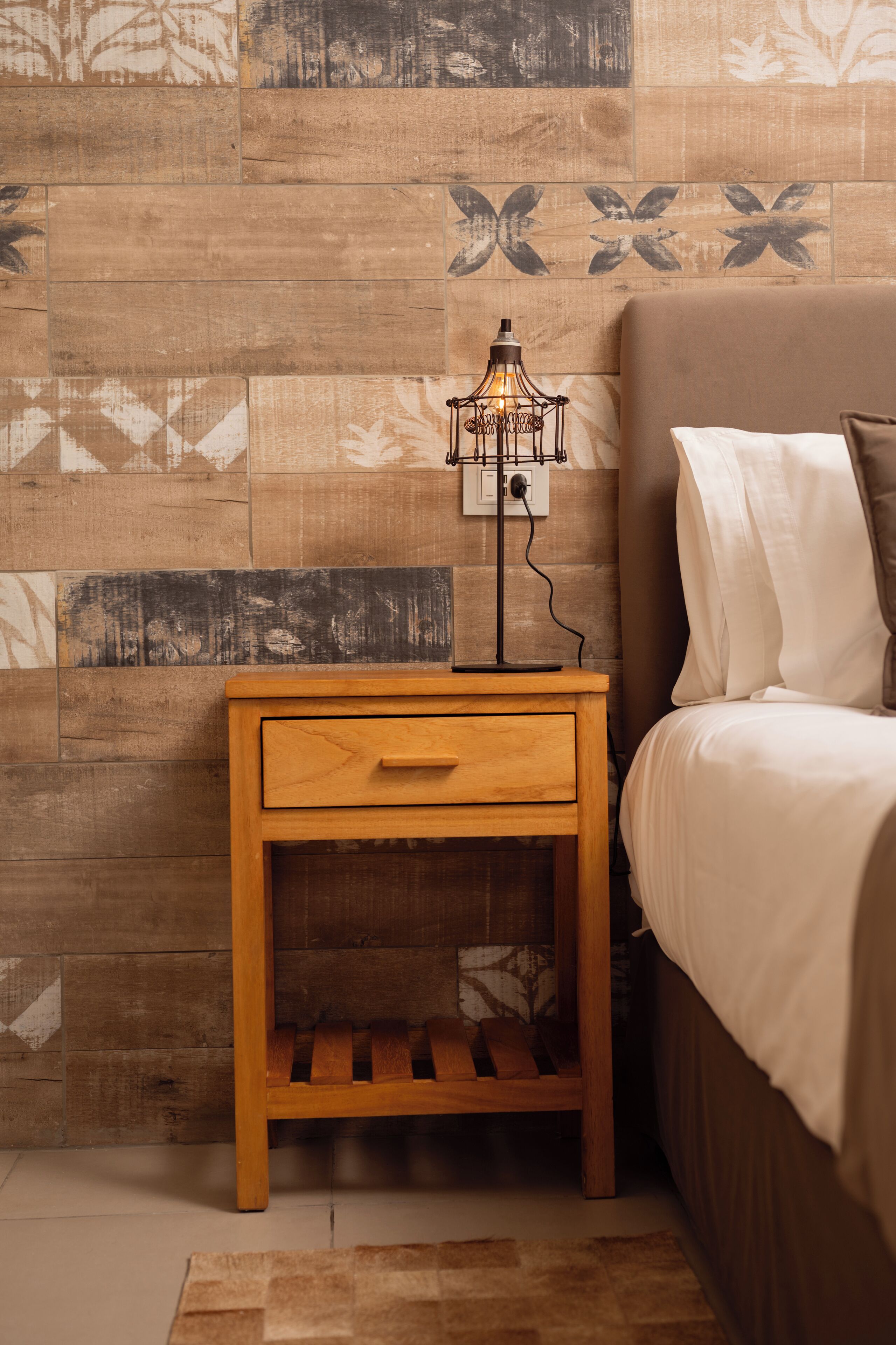 A bedroom with a wooden nightstand, a bedside lamp, a bed with white bedding, and a patterned wall.