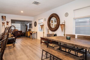 A spacious living room with a wooden dining table, benches, and a large mirror. There is a fireplace, a bookshelf, and a door leading to another room.
