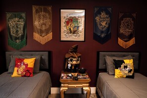A bedroom with two beds, each with themed pillows, a small table with books and a remote control, and a wall decorated with banners from the Harry Potter series.