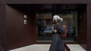 A modern building entrance with a large glass window, a visible car, and a person walking in front.