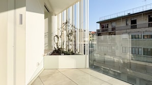 A modern balcony with a glass railing, a white planter with plants, and a view of neighboring buildings.