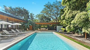 A rectangular swimming pool with lounge chairs and umbrellas on the deck, surrounded by trees and a covered seating area.