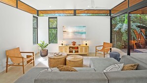 A modern living room with a grey sofa, two wooden chairs, a wooden cabinet, and a large window with a view of greenery.