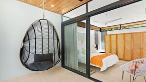 A modern bedroom with a hanging chair, a bed with an orange blanket, and a view through a glass door.