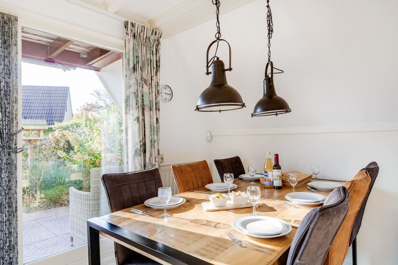 A dining area with a wooden table set for four, two hanging pendant lights, and a view of the garden through a window with curtains.