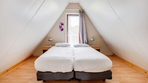 Una habitación con techo inclinado, una cama con dos almohadas, una mesita de noche y un ventanal con cortinas.