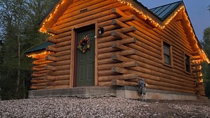 Une cabane en rondins avec une porte verte, entourée d’un chemin de gravier et d’arbres.
