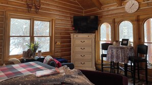 L’intérieur d’un chalet accueillant, avec des murs en bois, un téléviseur fixé au mur, une commode à tiroirs, une table recouverte d’une nappe à motifs floraux et une chaise.