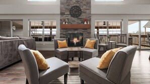 A spacious living room with a stone fireplace, grey sofas with yellow cushions, and large windows offering a view of the outdoors. - Lake Escape: Views, Hot Tub, Game Room & Pools (Hollister)