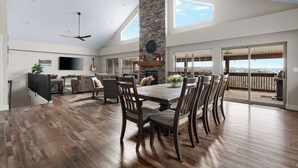 A spacious living area with a high ceiling, a fireplace, a dining table with chairs, and large windows offering a view of the outdoors. - Lake Escape: Views, Hot Tub, Game Room & Pools (Hollister)