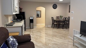 Una cocina moderna con armarios blancos, un refrigerador negro y un dispensador de agua. Hay un comedor con una mesa y sillas, y un televisor sobre un mueble.