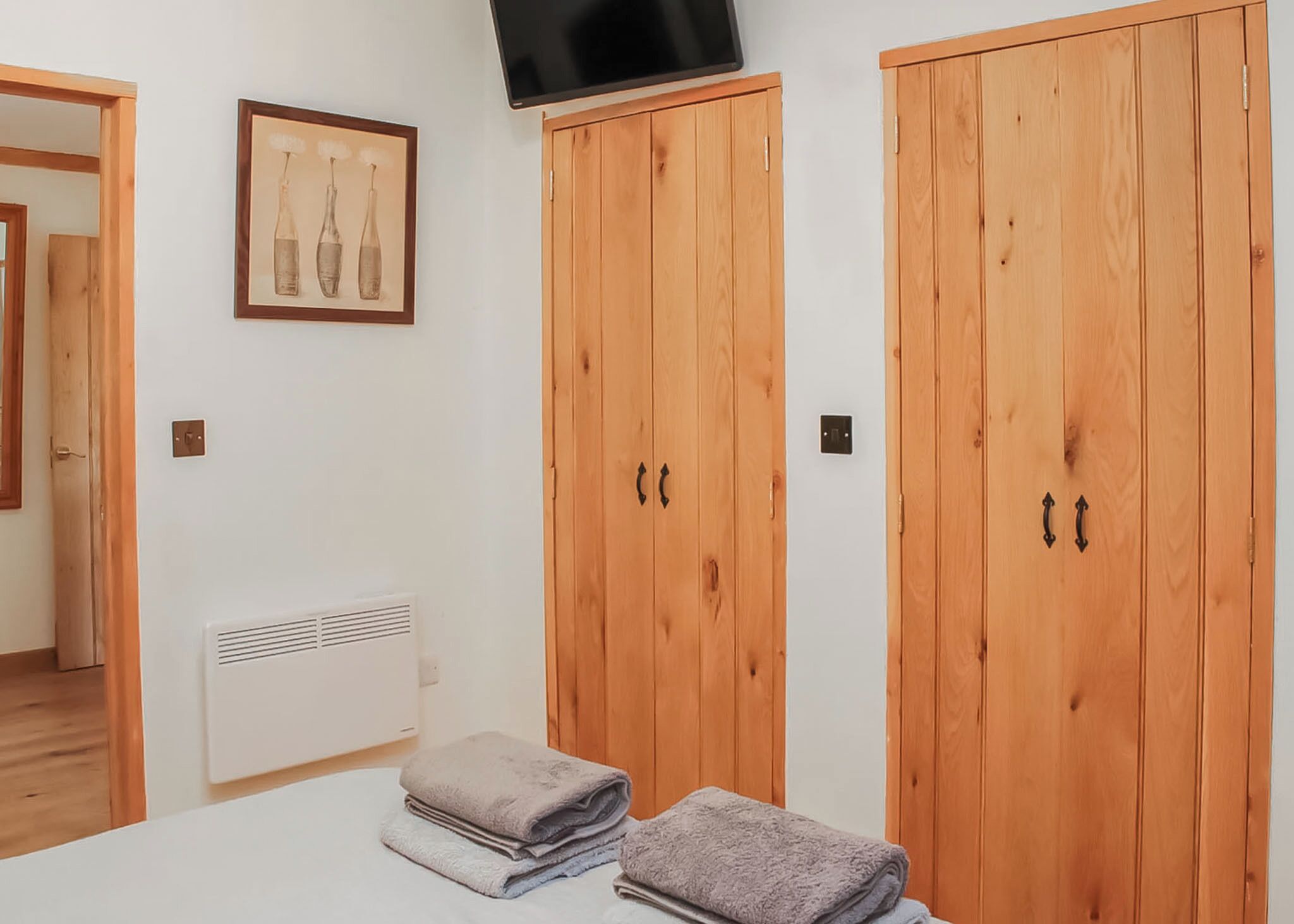 A bedroom with wooden wardrobes, a bed with folded towels, a wall-mounted TV, and a framed picture.