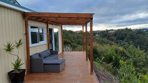 Ein überdachter Außenbereich mit Holzterrasse, grauem Sofa und Topfpflanzen.