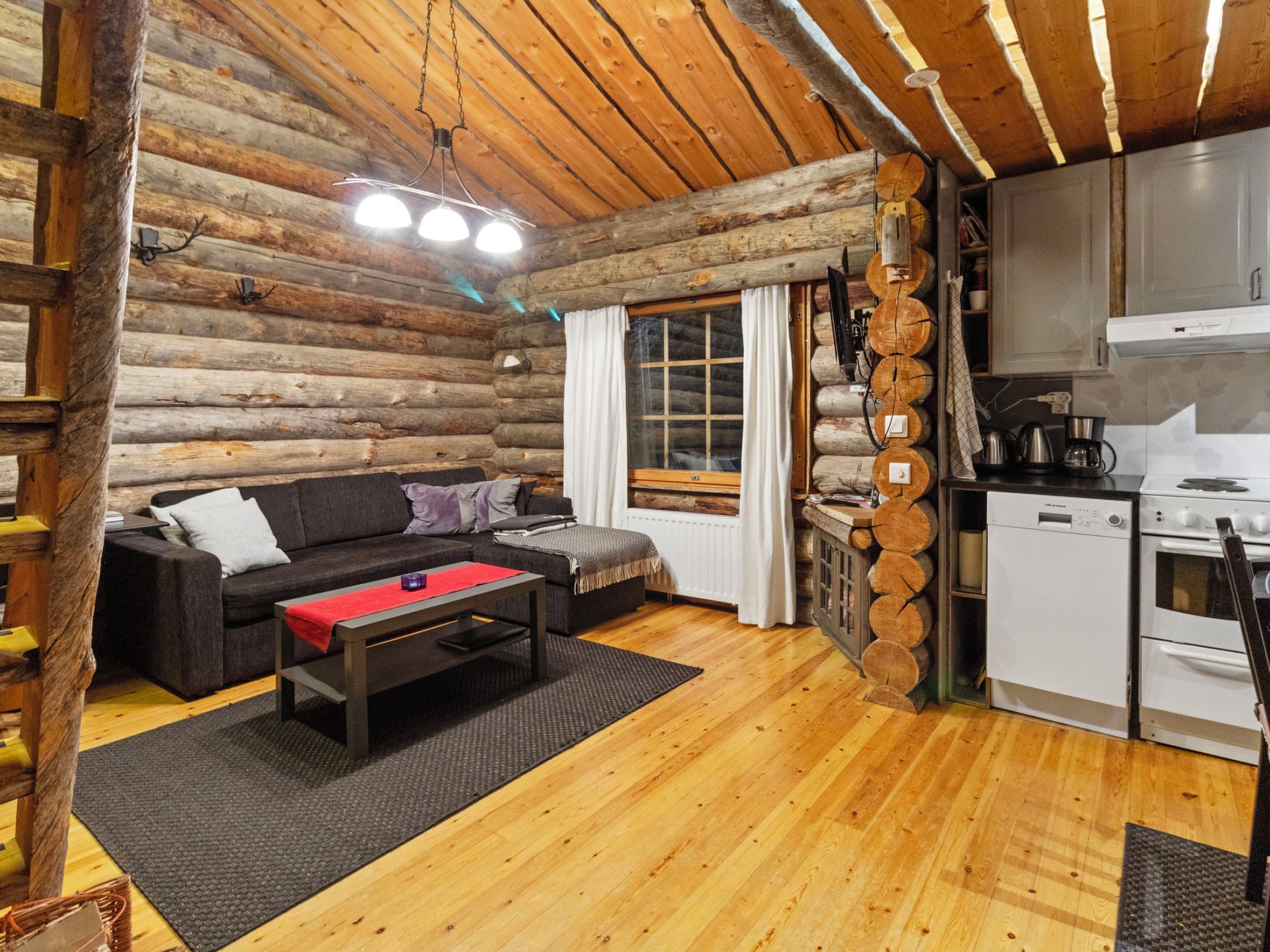 A log cabin interior with a sofa, a coffee table, a kitchen area with appliances, and a wooden ceiling.